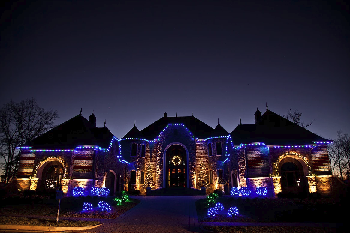 Large stone home outlined in blue and warm white Christmas lights at dusk