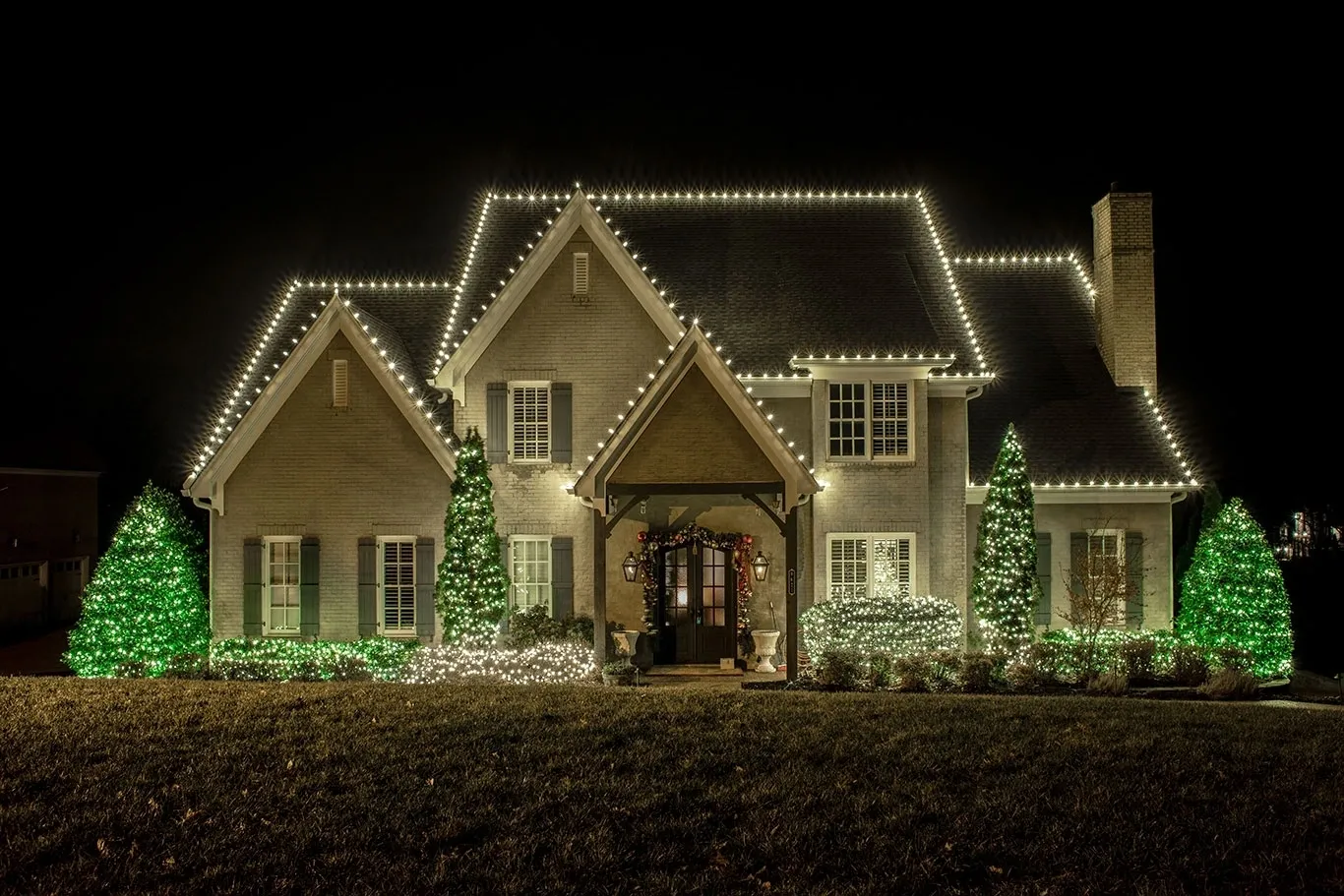 Home with white roofline Christmas lights and green-lit shrubs across the front