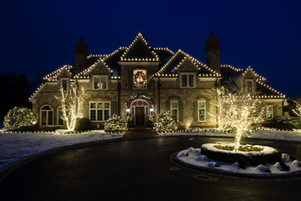 Stone home with warm white roofline lights and a lit tree beside the driveway at night