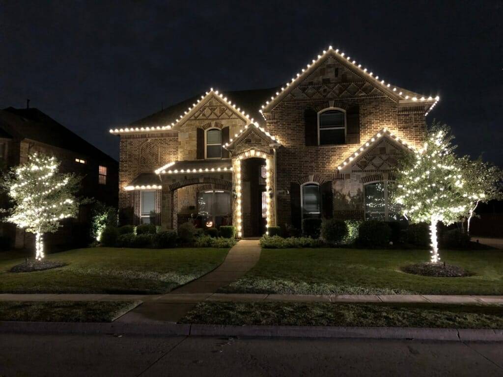 Brick two-story home with white roofline Christmas lights and two light-wrapped trees in the front yard