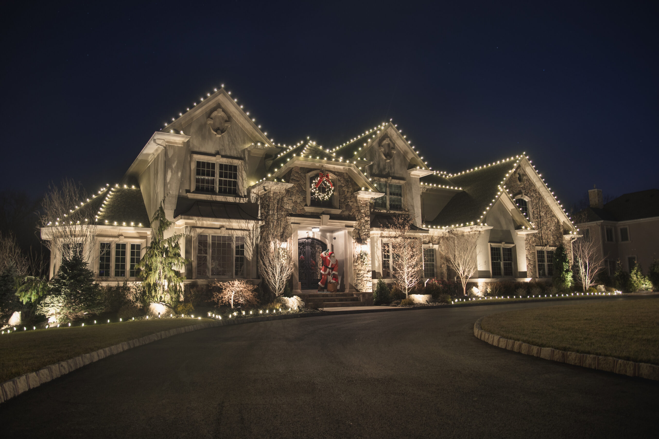 Large home with white roofline Christmas lights and lit trees at dusk